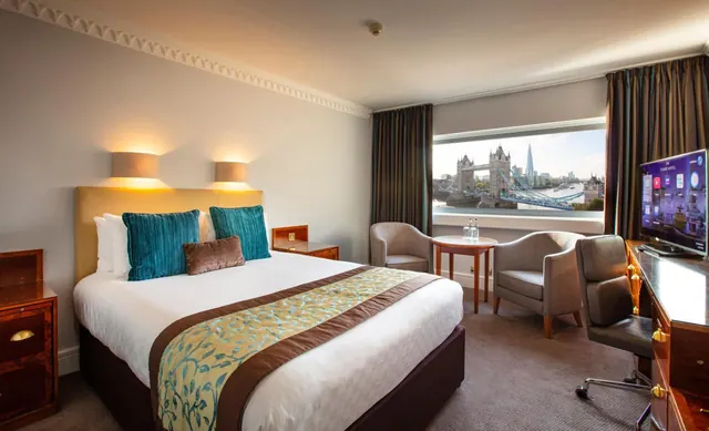 A hotel bedroom with a double bed, two armchairs, a desk and a television, with a window view of Tower Bridge and the Shard.