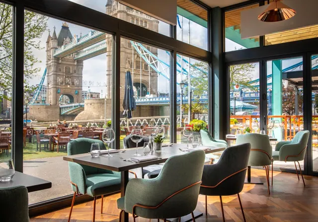 A restaurant dining room with floor-to-ceiling windows looking out over Tower Bridge and the River Thames, with set tables and green upholstered chairs.