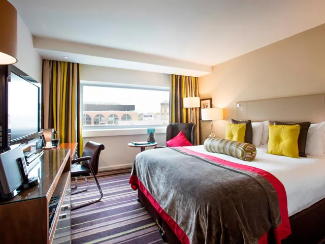 A staged hotel bedroom with a king bed, cushions, an armchair by the window, a desk with a television, and a view of a brick building outside.