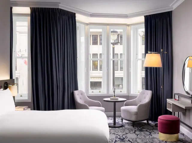 A bedroom with a large white bed, two armchairs beside a round side table, and a bay window with dark curtains looking onto buildings across the street.
