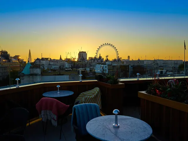A rooftop terrace with small marble tables and chairs looks out over London's skyline at sunset, with the London Eye visible.