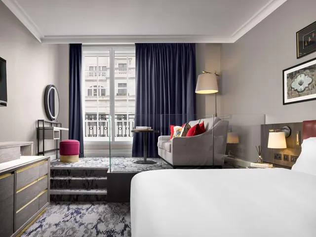 A hotel bedroom with a white king bed, a grey sofa with red cushions, and tall windows looking onto a building across the street.