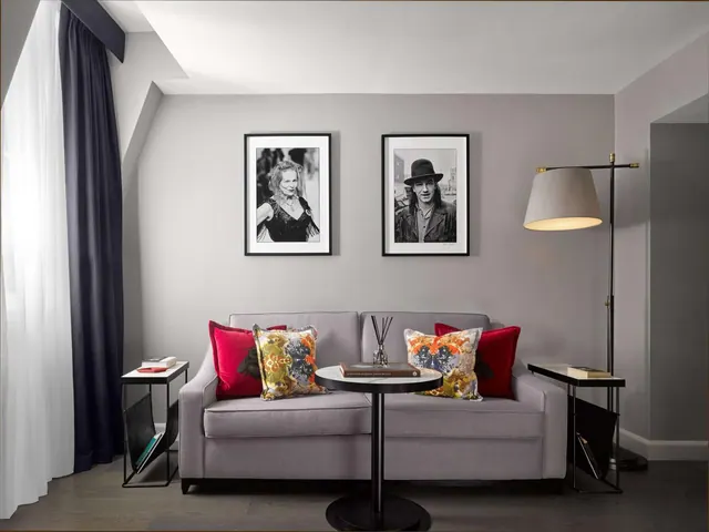 A living area with a grey sofa, red and patterned cushions, a round black side table, a floor lamp and two framed black and white portraits on the wall.