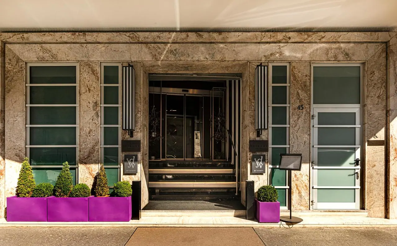 Front entrance of a marble-faced hotel building with glass doors, purple planters holding topiary shrubs, and a sign stand on the pavement.