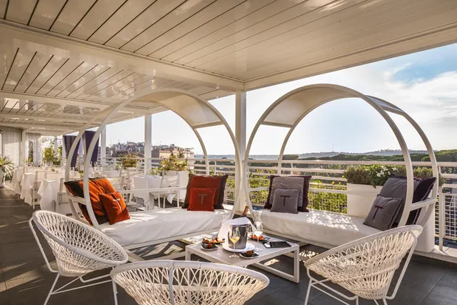 A rooftop terrace with white arched canopy daybeds, orange and grey cushions, wicker chairs and a low table holding glasses, a champagne bucket and small plates, with rooftops and trees beyond the railing.