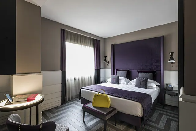 A bedroom with a large double bed under a tall purple upholstered headboard, a yellow handbag on a bench at the foot of the bed, and a window with sheer curtains.