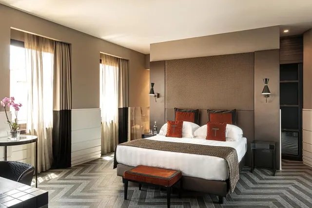 A double bed with orange cushions and a bench at its foot sits in a bedroom with tall curtained windows and chevron-patterned carpet.