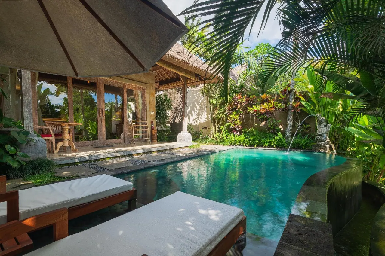 A small private pool with a water spout and sun loungers in the foreground, beside a thatched villa terrace and tropical garden.