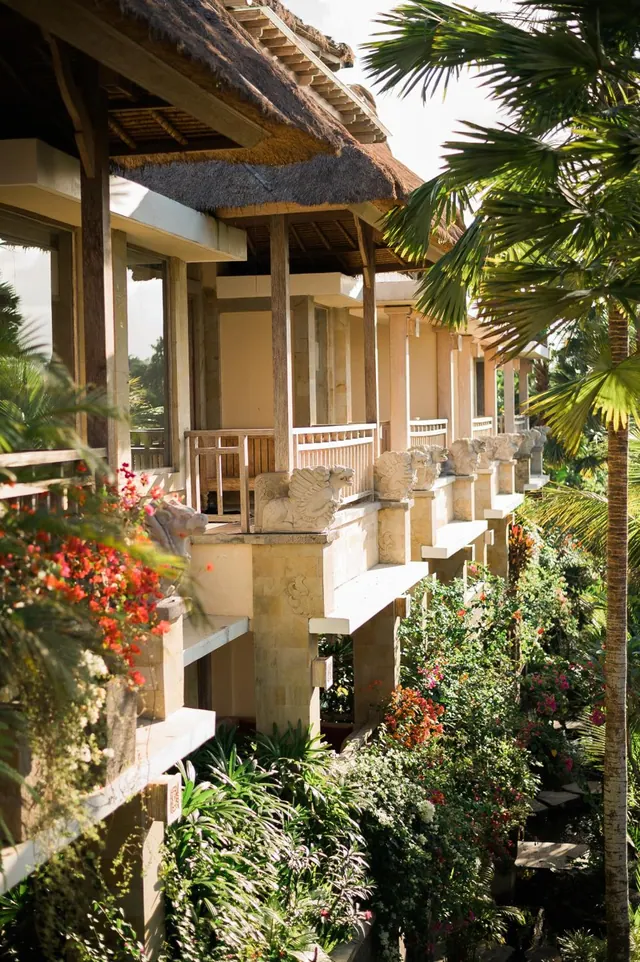 A Balinese-style building with a thatched roof, carved stone guardian figures on its balustrades, and tropical plants and a palm tree along its side.