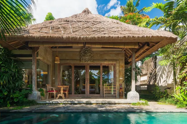 A thatched Balinese pavilion with carved wooden doors and a covered terrace with chairs, seen from the edge of a turquoise pool.