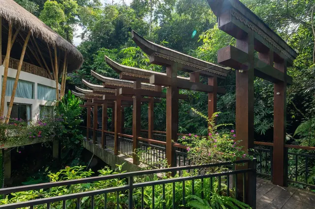 A covered wooden walkway with Balinese-style gates crosses a bridge over lush jungle, with a thatched building at the left edge.