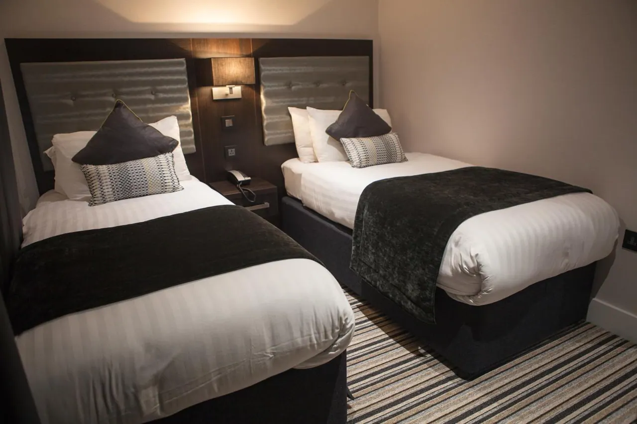 Two single beds with dark runners and striped white bedding sit against a dark wood headboard in a bedroom with striped carpet.