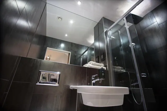 A modern bathroom with dark tiled walls, a semi-recessed white sink, folded towels on a shelf, and a glass-walled walk-in shower.