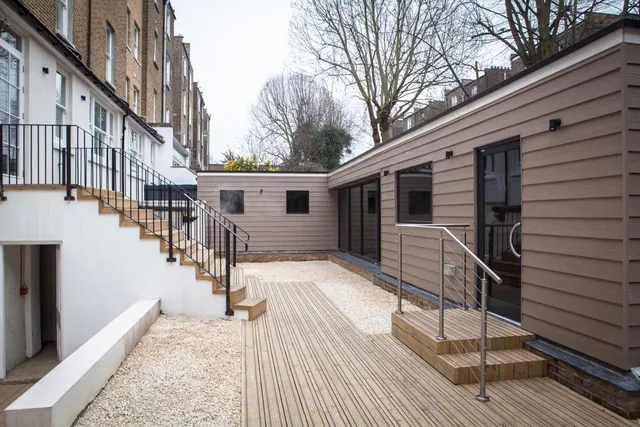 A rear courtyard with timber decking and gravel, an external staircase with metal railings, and a clad single-storey extension with glazed doors.