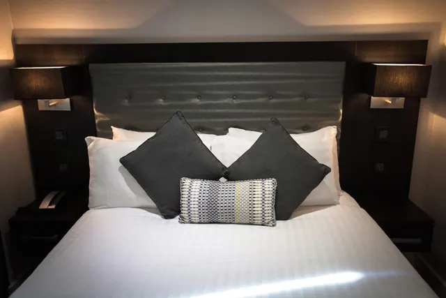 A made double bed with white linen, grey cushions and a patterned cushion against a dark upholstered headboard, flanked by wall lamps and bedside tables.