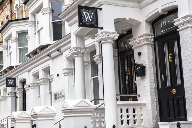 White painted townhouse facade with black hanging W signs, a black front door numbered 16-22, and a plaque reading The W Kensington beside the steps.