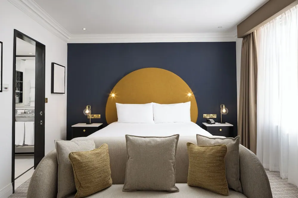 A double bed with a mustard circular headboard stands against a navy wall, with a curved grey sofa and cushions at the foot of the bed.