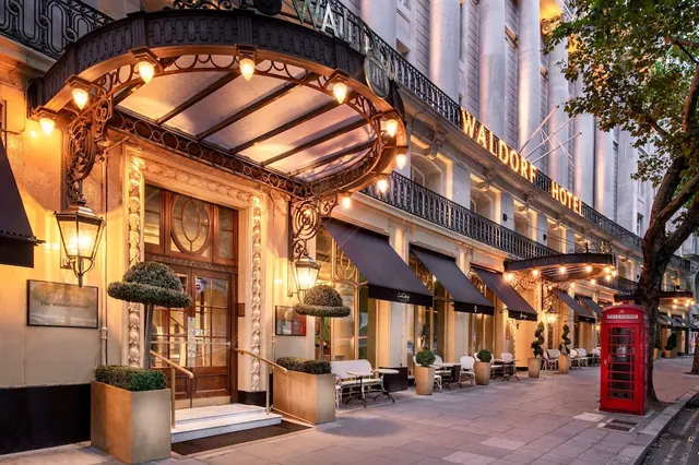 The Waldorf Hilton entrance facade at dusk, with a decorative canopy, lit lanterns, outdoor tables and chairs, and a red telephone box on the pavement.