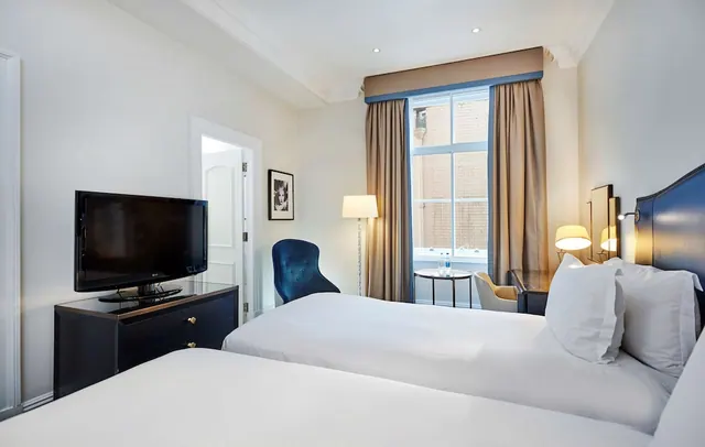 A bedroom with two beds, a flat-screen TV on a dark dresser, a blue armchair, and a window looking onto a brick building.