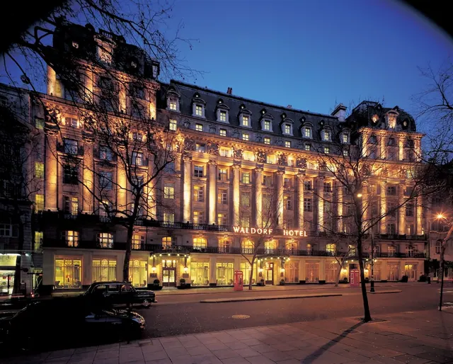 The illuminated facade of the Waldorf Hotel at dusk, with bare trees and parked cars along the street in front.