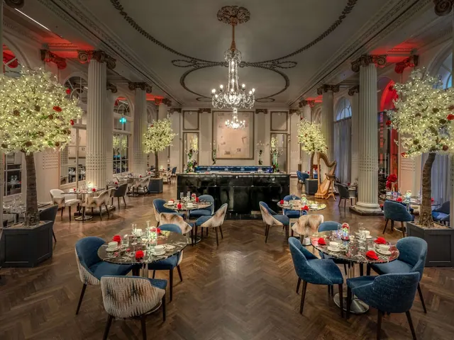 A dining room with round tables set with glassware and red napkins, blue upholstered chairs, a chandelier, illuminated trees and a harp in the background.