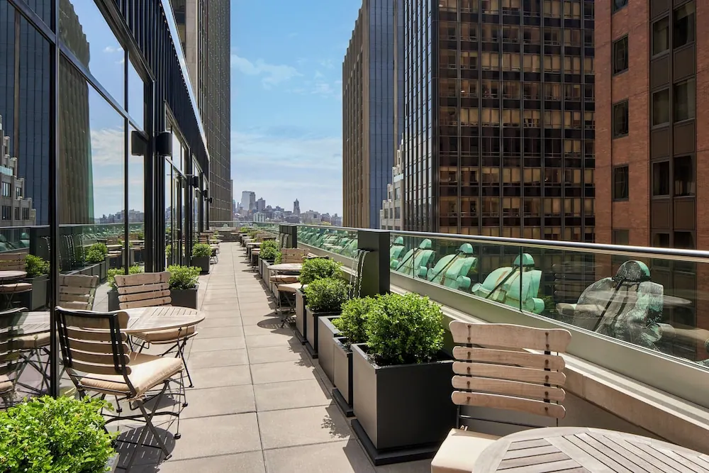 A rooftop terrace with wooden chairs, round tables, planters and a glass railing, flanked by tall buildings with a distant city skyline visible between them.