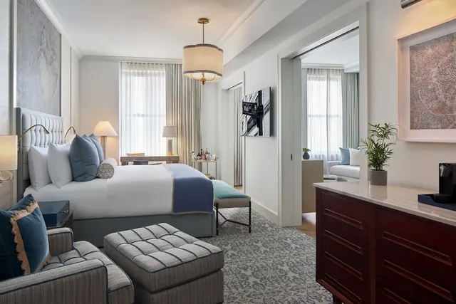 A staged hotel bedroom with a king bed, an upholstered armchair and ottoman, a wall-mounted television, and a sitting area visible through an open doorway.