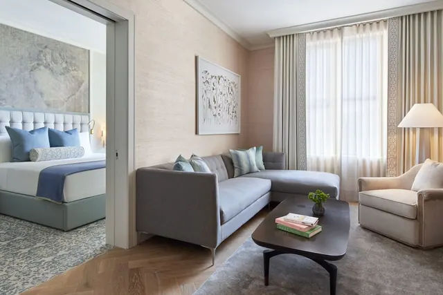 A living area with a grey sectional sofa, an armchair and a coffee table, with a bed visible through the doorway on the left.