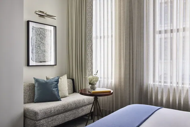 A bedroom corner with a grey upholstered sofa, blue and cream cushions, framed artwork, a round side table with flowers and books, sheer curtains over a window, and a bed edge in the foreground.