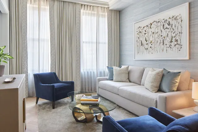 A living room with a grey sofa, two navy velvet armchairs, a glass coffee table with a gold base, and tall curtained windows.
