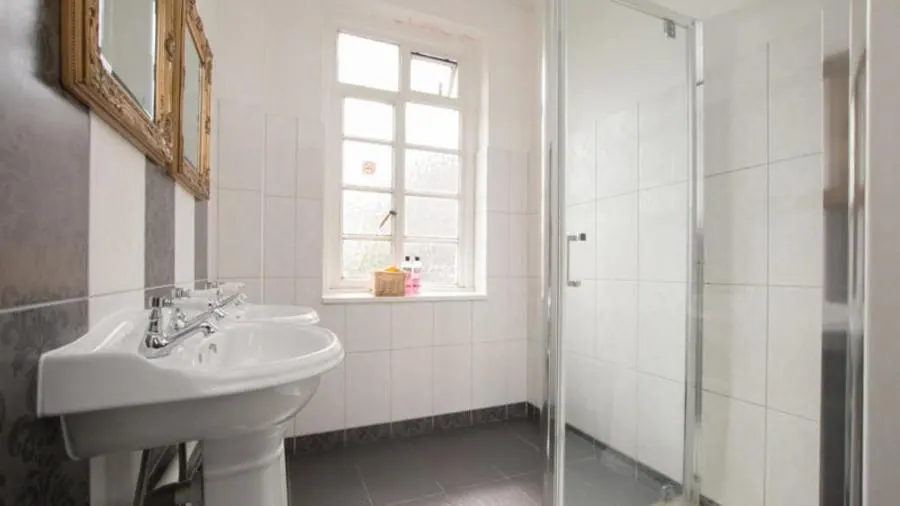 A bathroom with a double pedestal sink beneath ornate gold-framed mirrors, a glass-enclosed walk-in shower, and a window with toiletries on the sill.