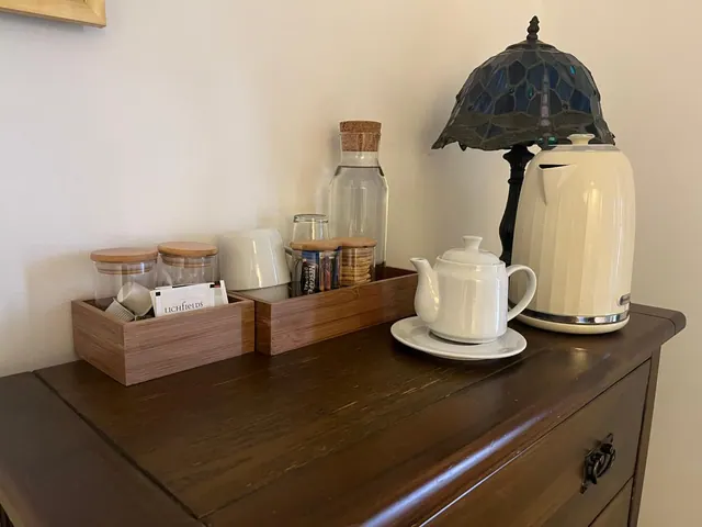 A tea and coffee station on a wooden dresser holds tea bags, instant coffee, biscuits, a water carafe, cups, a white teapot, a cream kettle and a stained-glass lamp.