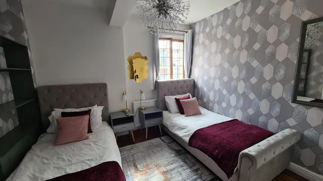 A bedroom with two single beds with grey tufted headboards, burgundy throws and pink cushions, beside a geometric patterned wallpaper wall and a window with grey curtains.