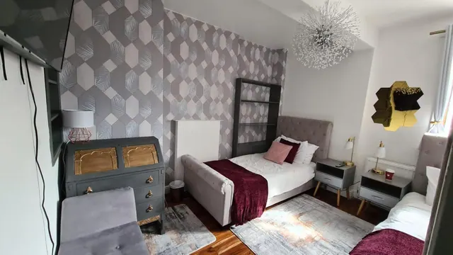 A bedroom with a double bed under a burgundy throw, a grey bureau with gold panels, bedside tables, a wall-mounted television and geometric grey wallpaper.