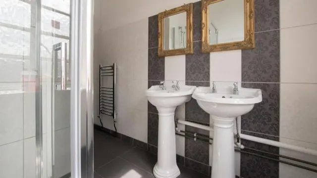 A bathroom with two white pedestal sinks, gold-framed mirrors above them, dark patterned tiles on the wall, and a glass shower enclosure on the left.