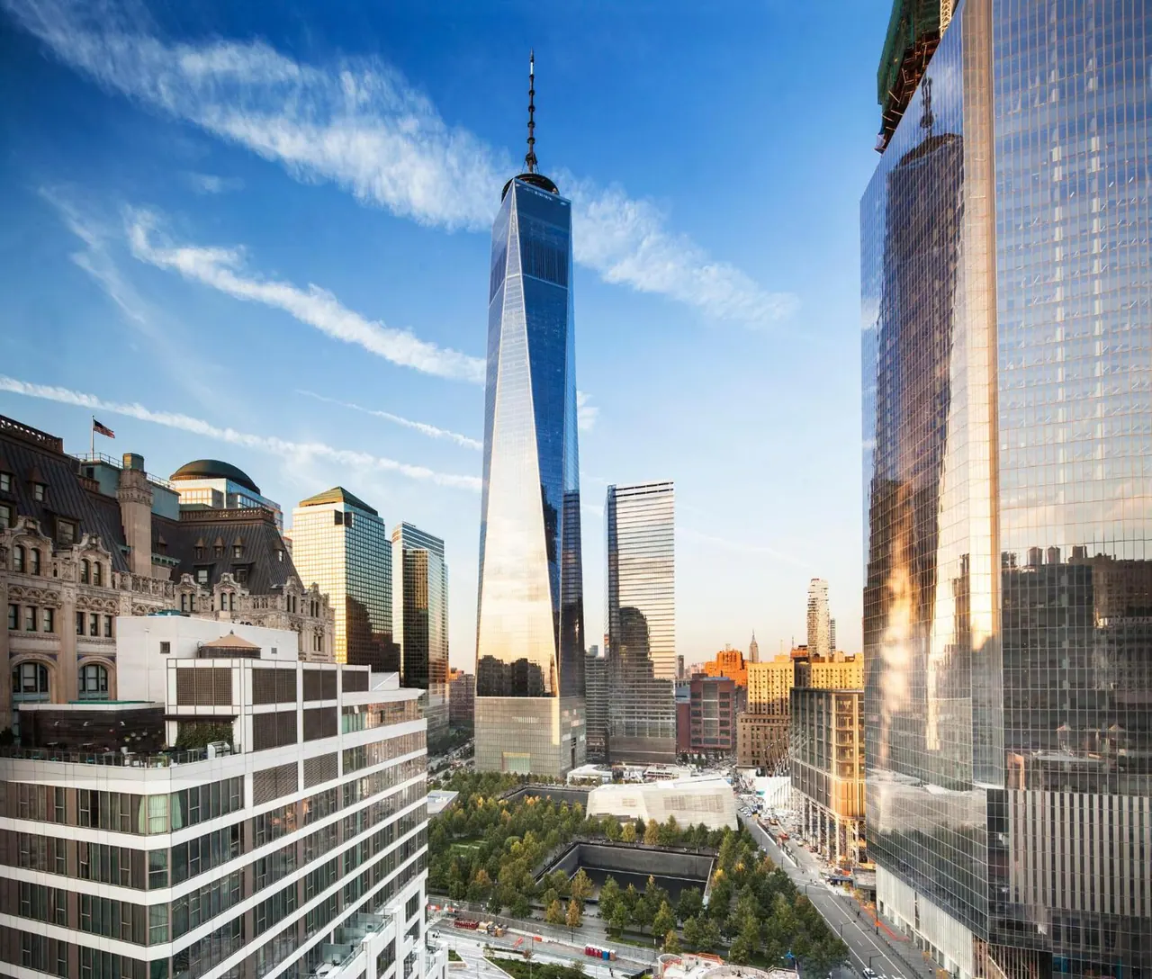View from an upper floor across surrounding office towers to One World Trade Center, with the 9/11 Memorial plaza and trees below.