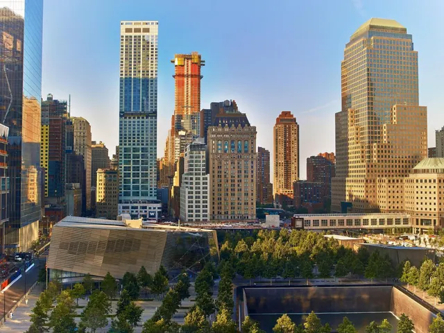 Elevated view across the National September 11 Memorial pool, trees and museum pavilion toward Lower Manhattan skyscrapers in warm late-day light.