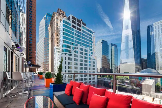 A rooftop terrace with a red cushioned sofa, high bar stools and a tall glass skyscraper rising above the surrounding city buildings.