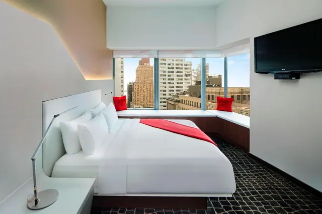 A king bed with a red throw and red cushions sits beside a window seat overlooking city buildings, with a wall-mounted television opposite.