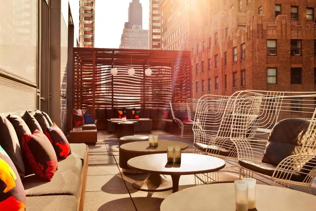 A rooftop terrace with grey sofas, wire chairs and round tables, with brick buildings and a distant spired skyscraper behind.