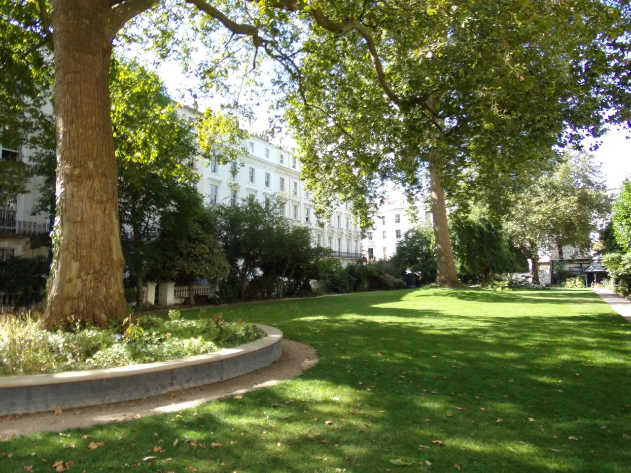 A lawned garden with large trees and a curved stone-edged flower bed, with white terraced buildings behind.