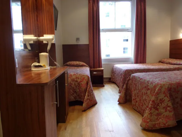 A hotel bedroom with two single beds under floral bedspreads, a wooden cabinet holding hairdryers and a kettle, and a window looking onto a neighbouring building.