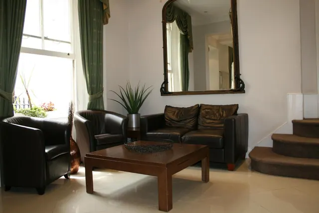 A lobby lounge with two leather armchairs, a leather sofa, a wooden coffee table, a large framed mirror, a potted plant and a carpeted staircase, with green curtains at the window.