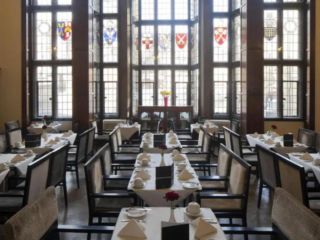 A formal dining room with white-clothed tables, upholstered chairs and tall leaded windows with heraldic stained glass.