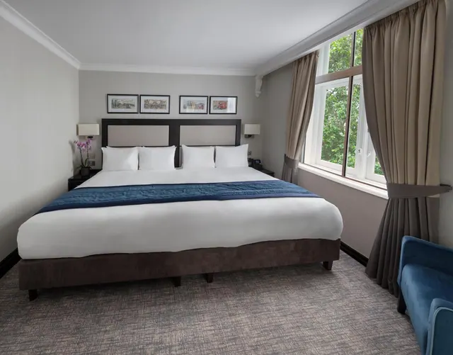 A bedroom with a king bed, bedside lamps, a blue armchair, and a window looking onto trees.
