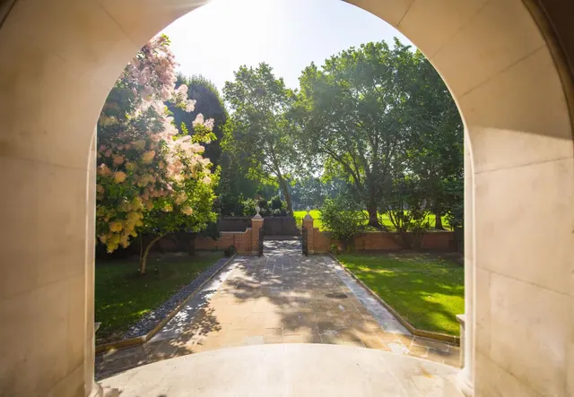 Looking through a stone archway onto a paved path that leads to a brick wall and gate, with lawns and mature trees beyond.