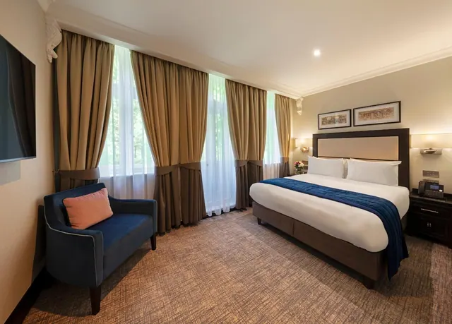 A made-up king bed with a navy throw sits beside a navy armchair and tall curtained windows that look out onto trees.