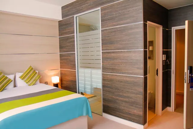 A bedroom with a made double bed under a green and teal striped throw, wood-panelled walls, and a glass-fronted shower enclosure and toilet area beside it.