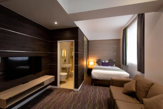 A hotel room with a wall-mounted television, a double bed with purple and grey cushions, a brown sofa, and an open doorway to a bathroom with a toilet and sink.
