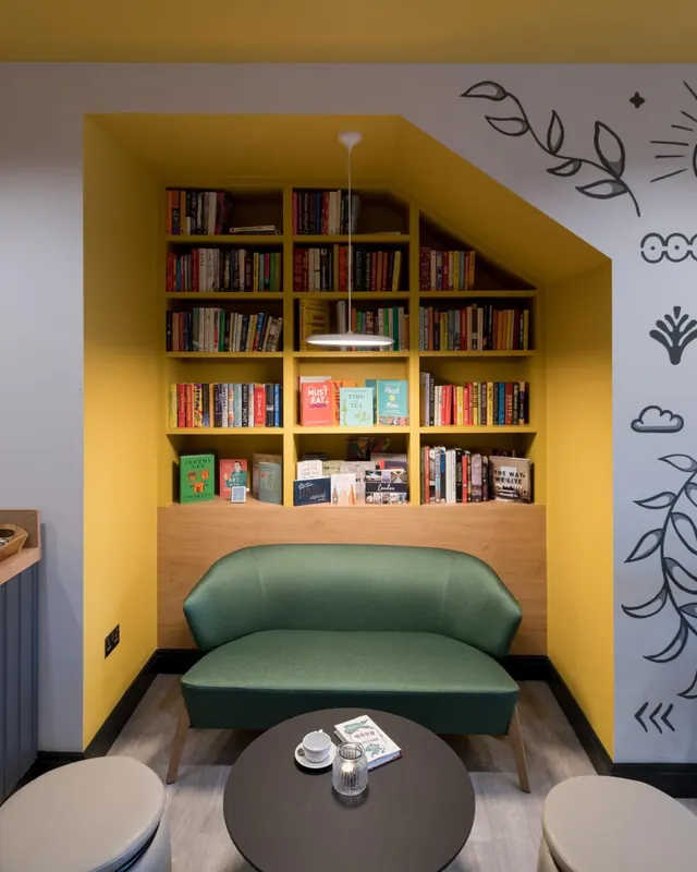 A reading nook with a yellow alcove of bookshelves, a green sofa, a round dark table holding a cup and a book, and two cream seats in the foreground.